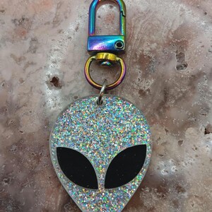 Op de afbeelding: Een glitterende alien sleutelhanger met een regenboogkleurige sluiting. De alienkop is zilverkleurig met zwarte ogen. De sleutelhanger is een leuk accessoire.