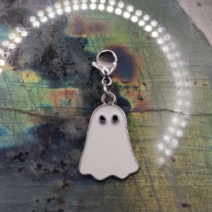 Op de afbeelding: Een zilverkleurige spookbedel met een witte emaille afwerking en zwarte ogen. De bedel heeft een karabijnsluiting voor bevestiging. De geest heeft een golvende onderrand. Perfect voor sieraden.