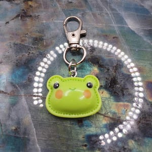 May include: A lime green frog-shaped keychain with a silver clasp. The frog has black eyes and rosy cheeks. The keychain is made of a soft, possibly rubbery material.