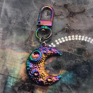 May include: A crescent moon keychain with a rainbow iridescent finish. The moon charm has a textured surface with small circular details and a large circular design. The keychain includes a lobster clasp and a small ring.
