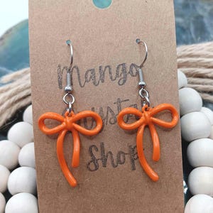 May include: A pair of orange bow earrings with silver hooks, displayed against a brown card with the text "Mango Crystal Shop". The earrings are a vibrant orange. The background features white wooden beads and rope.