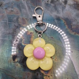 以下が含まれることがあります： 5枚の花びらを持つ黄色い花のチャームと、滑らかで丸いピンクの中心部が特徴のキーホルダー。チャームは小さな丸いリングで銀色の金属製ロブスタークラスプに取り付けられており、シンプルで機能的なアクセサリーとなっています。