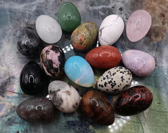Puede incluir: Una colección de huevos de piedras preciosas pulidas en varios colores, incluyendo verde, blanco, negro, rosa y moteado. Los huevos están dispuestos sobre una superficie marmolada, mostrando sus texturas y tonos únicos. Estos huevos decorativos son ideales para exhibir.