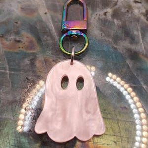 Puede incluir: Un dije con forma de fantasma de color rosa claro hecho de un material liso y veteado. El dije presenta dos aberturas ovaladas para los ojos y está sujeto a un broche de langosta metálico con acabado iridiscente de arcoíris.