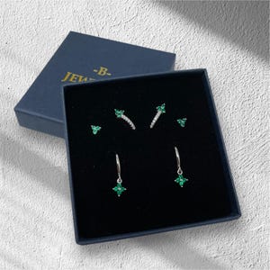 May include: A set of five silver earrings with green gemstones. The earrings are displayed in a black velvet box with a blue lid. The lid has the text "B-Jew" printed on it.