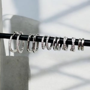 May include: A selection of silver hoop earrings, presented on a black bar. The earrings feature various designs and sizes, including some with clear gemstones. The earrings are arranged in a line, displaying different styles.