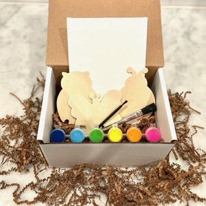 May include: A white box filled with a paint-your-own craft kit. The kit includes three wooden shapes: a butterfly, a ladybug, and an owl. The box also contains six small paint pots in various colors, a paintbrush, and a blank canvas.