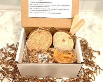 May include: A white cardboard box with a brown lid, containing a bird feeder kit. The kit includes two wooden circles, a small container of peanut butter, and a bag of birdseed. The box lid has instructions for making the bird feeder, which include a warning that the kit contains peanut products.