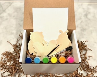 May include: A white box filled with a paint-your-own craft kit. The kit includes three wooden shapes: a butterfly, a ladybug, and an owl. The box also contains six small paint pots in various colors, a paintbrush, and a blank canvas.