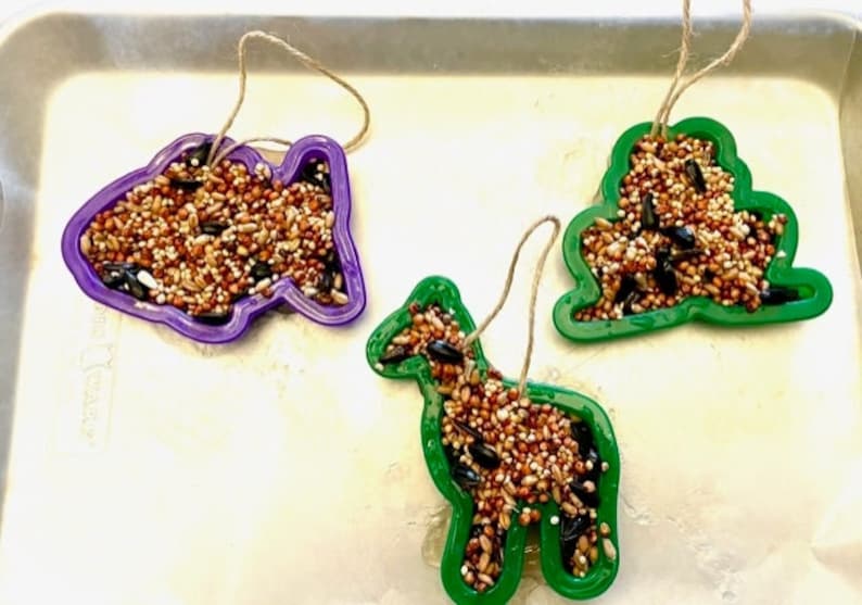May include: Three bird feeders made from cookie cutters filled with birdseed. The feeders are shaped like a fish, a tree, and a horse. The feeders are purple, green, and green. Each feeder has a string attached to it.