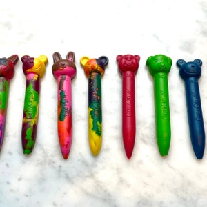 May include: Ten colorful crayons with animal-shaped tops. The crayons are arranged in a row on a white surface. The crayons are all different colors, including yellow, pink, green, brown, red, blue, and purple. The animal shapes include a lion, a cow, a bear, a rabbit, a mouse, a dog, and a cat.
