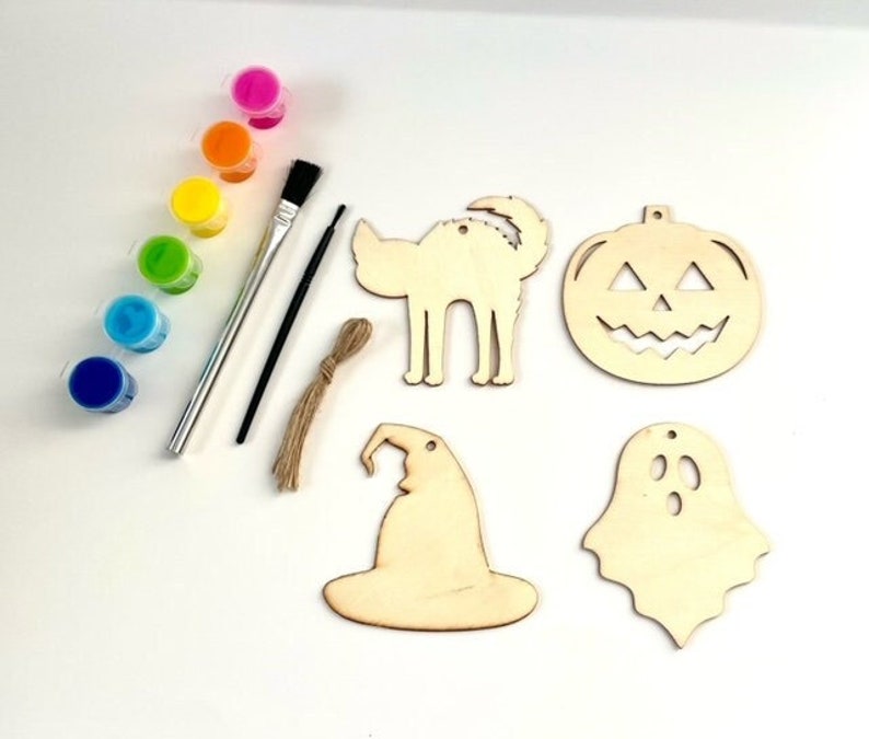 May include: A Halloween craft kit with six wooden ornaments, a paintbrush, and six small bottles of paint. The ornaments are shaped like a pumpkin, a ghost, a witch's hat, a cat, and two bats. The paint is in rainbow colors.