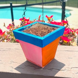 May include: A small, square-shaped planter with a blue, pink, and orange paint design. The planter is filled with brown soil.