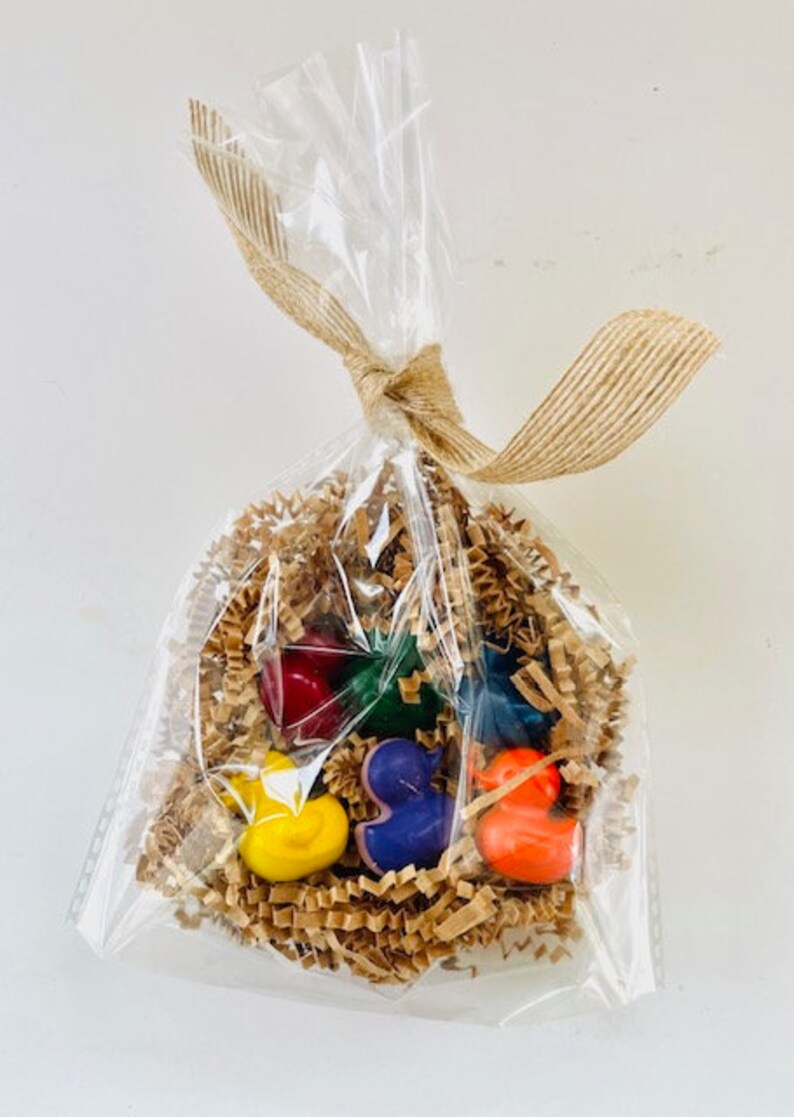 May include: A clear plastic bag filled with brown crinkle paper and six colorful crayon shaped like ducks. The crayons are red, yellow, green, blue, orange, and purple.