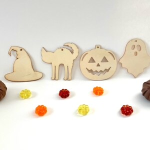 May include: Five wooden Halloween ornaments, including a witch's hat, a cat, a pumpkin, and a ghost. The ornaments are surrounded by small, colorful candies.
