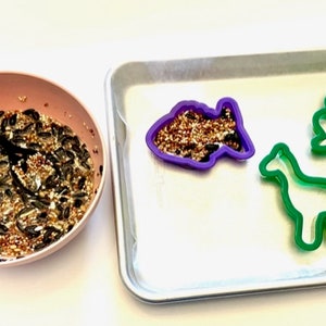 May include: A pink bowl filled with birdseed, a baking sheet with three cookie cutters: a purple fish, a green giraffe, and a green cloud.