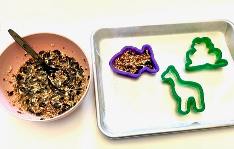May include: A pink bowl filled with birdseed, a baking sheet with three cookie cutters: a purple fish, a green giraffe, and a green cloud.
