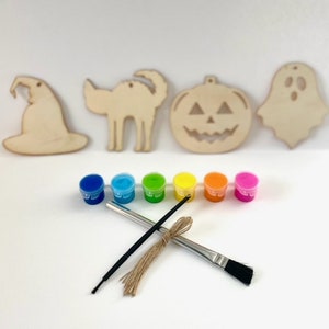 May include: A set of five unfinished wooden Halloween ornaments, including a witch's hat, a cat, a pumpkin, and a ghost. The ornaments are ready to be painted with the included set of six paint pots and a paintbrush. The paint pots are arranged in a rainbow order from blue to pink.