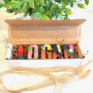 May include: A cardboard box with the name "LOGAN" spelled out in colorful crayon letters. The letters are sitting on brown packing paper inside the box. The box is open and there is a brown ribbon tied around it.