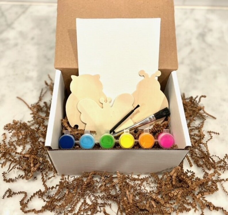 May include: A white box filled with a paint-your-own craft kit. The kit includes three wooden shapes: a butterfly, a ladybug, and an owl. The box also contains six small paint pots in various colors, a paintbrush, and a blank canvas.