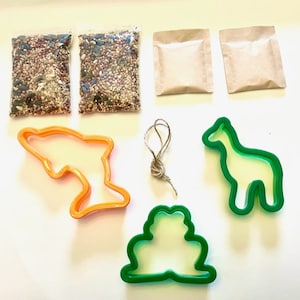 May include: A craft kit for making bird feeders includes two bags of birdseed, two brown paper bags, a green frog cookie cutter, a green giraffe cookie cutter, an orange dolphin cookie cutter, and a length of twine.