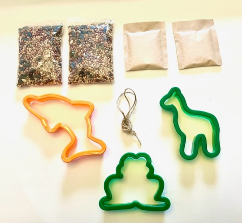 May include: A craft kit for making bird feeders includes two bags of birdseed, two brown paper bags, a green frog cookie cutter, a green giraffe cookie cutter, an orange dolphin cookie cutter, and a length of twine.