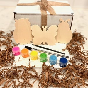 May include: A craft kit with three wooden shapes: an owl, a butterfly, and a ladybug. The kit includes five small paint pots in orange, yellow, green, blue, and light blue, and two paintbrushes. The kit is packaged in a white box with a brown ribbon.