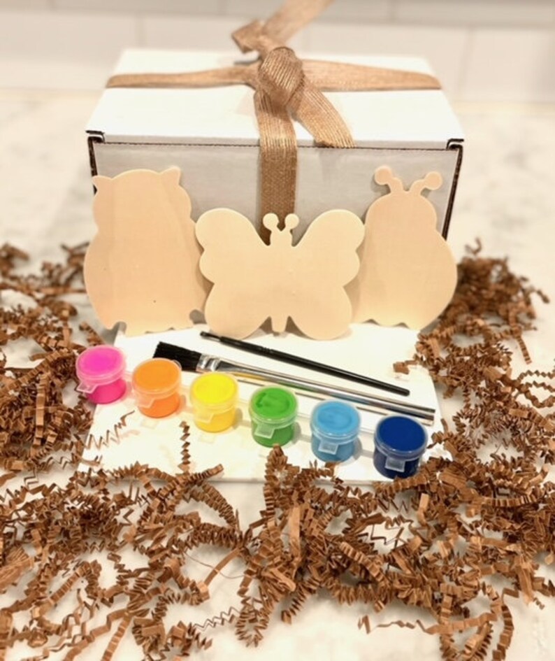 May include: A craft kit with three wooden shapes: an owl, a butterfly, and a ladybug. The kit includes five small paint pots in orange, yellow, green, blue, and light blue, and two paintbrushes. The kit is packaged in a white box with a brown ribbon.