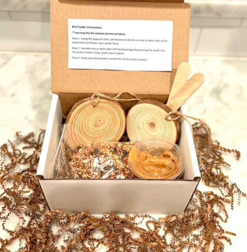 K&ouml;nnte beinhalten: Eine wei&szlig;e Pappbox mit braunem Deckel, die ein Vogelh&auml;uschen-Set enth&auml;lt. Das Set beinhaltet zwei Holzscheiben, einen kleinen Beh&auml;lter mit Erdnussbutter und eine T&uuml;te Vogelfutter. Der Boxdeckel enth&auml;lt Anweisungen zum Bau des Vogelh&auml;uschens, die einen Warnhinweis enthalten, dass das Set Erdnussprodukte enth&auml;lt.