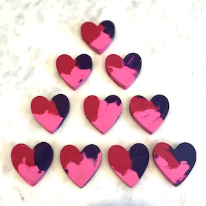May include: Nine heart-shaped crayons in shades of pink and purple. The crayons are arranged in a triangle shape.
