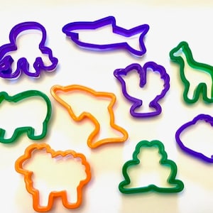 May include: A set of ten colorful plastic cookie cutters shaped like various animals and sea creatures. The cutters are purple, green, and orange. The shapes include an octopus, a shark, a giraffe, a cactus, a dolphin, a hippopotamus, a sheep, and a fish.