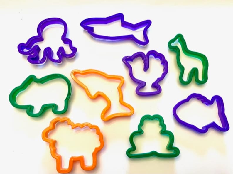 May include: A set of ten colorful plastic cookie cutters shaped like various animals and sea creatures. The cutters are purple, green, and orange. The shapes include an octopus, a shark, a giraffe, a cactus, a dolphin, a hippopotamus, a sheep, and a fish.