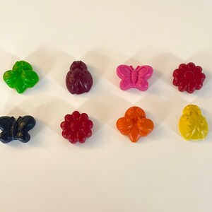May include: Eight colorful wax crayons shaped like a ladybug, butterfly, and flower. The crayons are arranged in a row on a white surface.