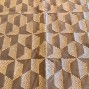 Puede incluir: Primer plano de una tela beige con un patrón texturizado de cubos 3D. La tela tiene un brillo sutil y una sensación suave y mullida.
