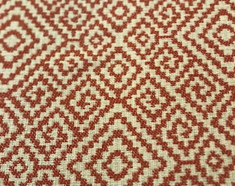 May include: A close-up of a fabric with a repeating geometric pattern. The pattern is made up of small squares and diamonds in a rust orange colour on a cream background.