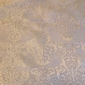 Op de afbeelding: Een witte stof met een gouden damastpatroon. Het patroon is een herhalend ontwerp van spiralen en bladeren.