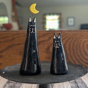 May include: Two black ceramic cat figurines with white facial details and a yellow crescent moon. The taller cat has a moon on its head. The cats are on a decorative black metal stand.
