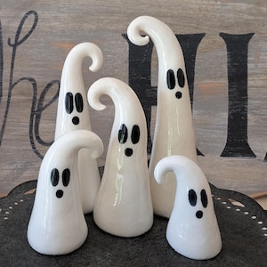 May include: Five white ceramic ghost figurines with black eyes and mouths. The ghosts have a glossy finish and varying heights, with curved tops. The figurines are arranged on a dark surface, perfect for Halloween decor.