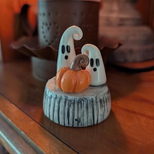 May include: A ceramic figurine featuring two white ghosts with black eyes and mouths standing behind a small orange pumpkin with a brown stem. The figures are mounted on a round, textured base with a weathered, grey, wood-like finish.
