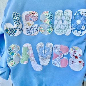 May include: Light blue sweatshirt with the words "JESUS SAVES" appliqued on the back. The letters are made from various floral and patterned fabrics, creating a patchwork design. The sweatshirt has long sleeves.