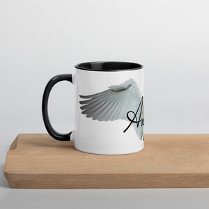 May include: White ceramic mug with a black interior and handle. The mug features a design of a white angel wing with the word "Angel" in a script font. The mug is sitting on a wooden cutting board.
