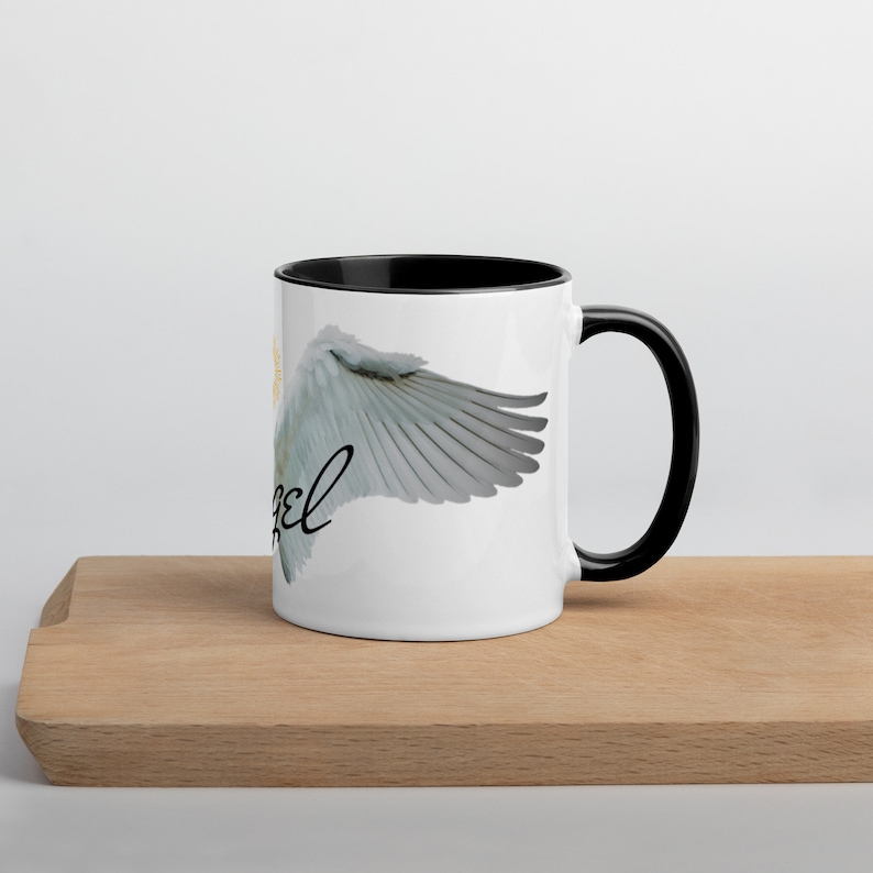 May include: White ceramic mug with a black interior and handle. The mug features a design of angel wings and the word "Angel" in a script font. The wings are light gray with detailed feathering, and a gold halo is visible.