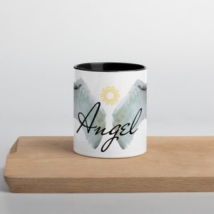 May include: White ceramic mug with a black interior, featuring a design of angel wings and the word "Angel" in a script font. A gold sunburst design is between the wings. The mug is on a wooden surface.