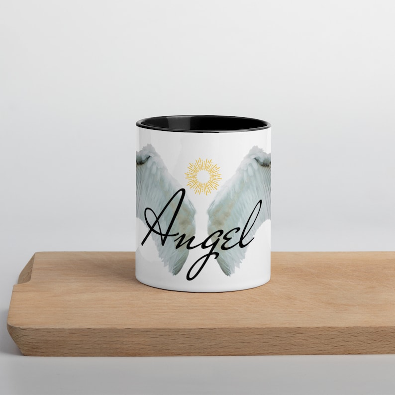 May include: White ceramic mug with a black interior, featuring a design of angel wings and the word "Angel" in a script font. A gold sunburst design is between the wings. The mug is on a wooden surface.