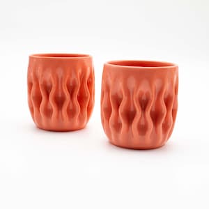 May include: Two coral-colored ceramic cups with a wavy, textured design. The cups have a cylindrical shape and are set against a white background. The cups are approximately 3.5 inches tall.