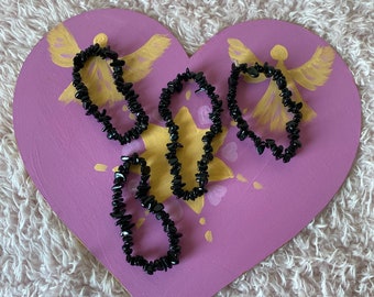 May include: Three black chip bracelets on a pink heart-shaped background with gold painted angel wings and a star.