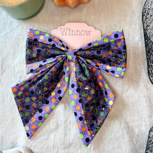 May include: A large hair bow featuring a purple fabric base with a polka dot pattern in orange, lime green, and black. The bow is layered with a black glittery mesh overlay and is attached to a light pink display card with the text Winnow printed.