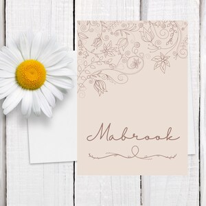 以下が含まれることがあります： 黄色い中心を持つ白いデイジーが、ベージュ色のカードの隣にある白い木の表面に置かれています。カードには花の線画と、下部に装飾的な線が付いたスクリプトフォントで「Mabrook」という名前が描かれています。