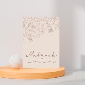 以下が含まれることがあります： フローラルな線画と筆記体フォントで「Mabrook」という名前が入ったベージュ色のカード。カードは木製のスタンドに大理石の球体と一緒に飾られています。フローラルなデザインは右上隅にあります。