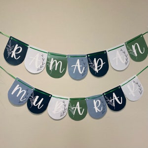 May include: A banner with the word "RAMADAN MUBARAK" spelled out on a string of colorful pennants. The pennants are in shades of blue, green, and white, with floral accents and white lettering. The banner is designed for festive occasions.